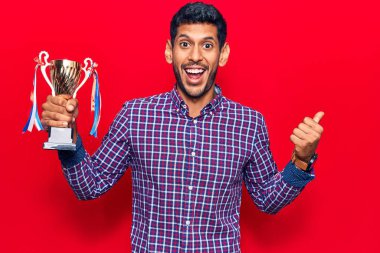 Genç latin adam elinde kupa tutarken başparmağını yana kaldırıyor ve ağzı açık gülümsüyor. 