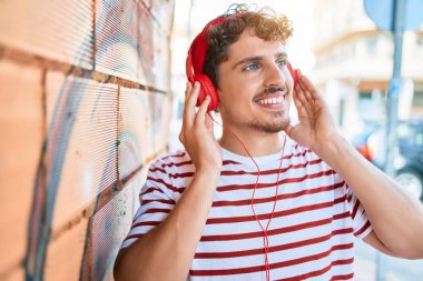 Genç beyaz adam gülümsüyor ve şehirde kulaklıkla müzik dinliyor..