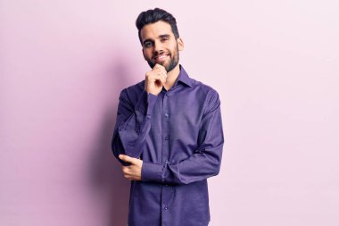 Sakallı, rahat gömlekli, kameraya kendinden emin, kolları çapraz ve elleri çenesinde gülümseyen genç bir adam. Olumlu düşünmek. 