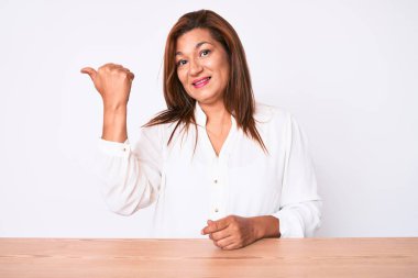 Orta yaşlı esmer, İspanyol iş kadını. Masada oturan, gülümseyen, gülümseyen ve baş parmağıyla yüzünü gösteren.. 