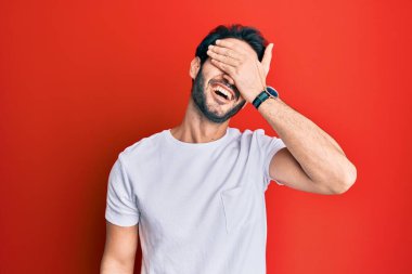 Latin asıllı genç bir adam, günlük beyaz tişört giyiyor, gülümsüyor ve elleri yüzünde, şaşırtmak için gözleri kapatıyordu. kör kavram. 