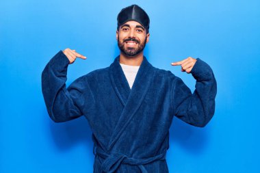 Genç İspanyol adam uyku maskesi takıyor ve bornoz giyiyor. Yüzünde bir gülümsemeyle, kendini gururlu ve mutlu bir şekilde gösteriyor.. 