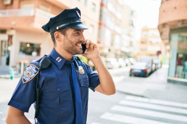 Genç, yakışıklı İspanyol polis memuru polis üniforması giyip mutlu gülümsüyor. Yüzünde tebessümle dikilip şehrin caddesinde akıllı telefondan konuşuyor..