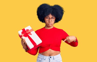 Genç Afro-Amerikalı kız elinde hediyeyle üzgün ve üzgün görünüyor, parmaklarıyla yönü gösteriyor, mutsuz ve depresif.. 