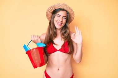 Güzel beyaz kadın, yaz küreği ve kova oyuncaklarıyla oynuyor. Parmaklarıyla imza atıyor, gülümsüyor. 