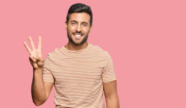 Yakışıklı, günlük kıyafetler giyen, üç numaralı parmaklarıyla gösteriş yapan, kendinden emin ve mutlu gülümseyen İspanyol.. 