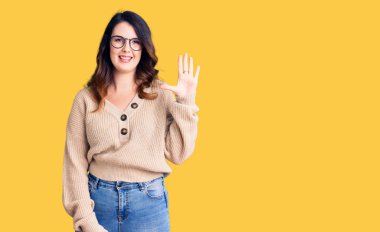 Güzel, genç, esmer bir kadın. Günlük kıyafetler ve gözlük takıyor. Beş numaralı parmaklarıyla gösteriyor ve işaret ediyor.. 