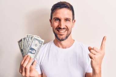 Soyutlanmış beyaz bir zemin üzerinde bir deste para tutan yakışıklı genç adam mutlu bir şekilde elini ve parmağını kenarı işaret ediyor.