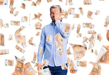 Zarif bir gömlek giyen yakışıklı genç bir adam bir gözünü eliyle kapatıyor, yüzünde kendine güvenen bir gülümseme ve sürpriz bir duygu var..