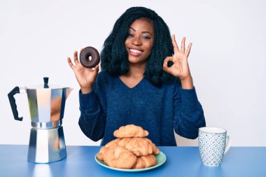 Güzel bir Afrikalı kadın kahvaltısını yapıyor çikolatalı donut tutuyor tamam işareti yapıyor, gülümsüyor dostça bir jest yapıyor mükemmel bir sembol. 