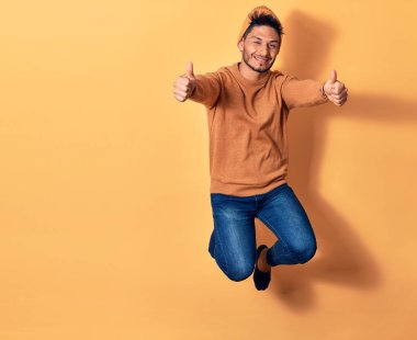 Gündelik kıyafetler giyen ve yün şapka takan genç yakışıklı Latin adam mutlu gülümsüyor. Yüzünde tebessümle zıplıyor, tamam işareti yapıyor, sarı arka plan üzerinde başparmaklar var.