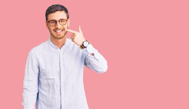 Zarif bir iş gömleği ve gözlüklü yakışıklı genç adam. Yüzüne ve burnuna parmak gösteren, neşeli gülümseyen. güzellik konsepti 