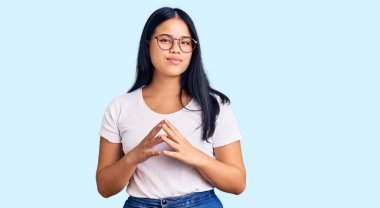 Genç ve güzel Asyalı bir kız günlük kıyafetler ve gözlük takıyor ellerini birleştirip gülümsüyor ve rahatlıyor. Başarı ve iyimserlik 