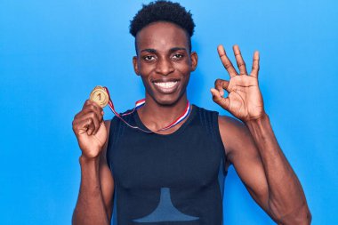 Afro-Amerikalı genç adam spor giysisi takıyor elinde madalyayla tamam işareti yapıyor, gülümseyerek mükemmel bir sembol oluşturuyor. 