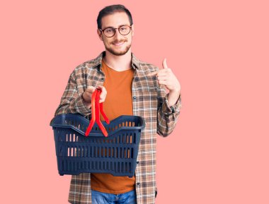Genç, beyaz bir adam elinde süpermarket alışveriş sepeti tutarak mutlu ve gururlu bir kişiyi işaret ediyor. 