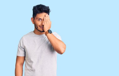 Yakışıklı Latin Amerikalı genç adam gündelik tişört giyiyor bir gözünü eliyle kapatıyor, yüzünde kendine güvenen bir gülümseme ve sürpriz bir duygu var.. 