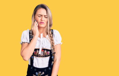Oktoberfest elbisesi giyen genç, sarışın bir kadın diş ağrısı ya da diş hastalığından dolayı acı dolu bir ifadeyle dudaklarına dokunuyor. dişçi 
