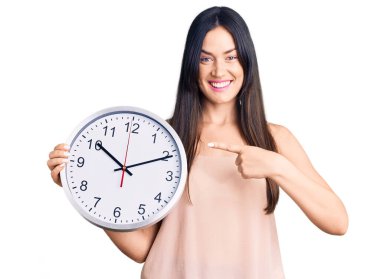 Genç, güzel, beyaz bir kadın büyük bir saat tutuyor. Gülümsüyor, mutlu bir şekilde işaret ediyor. 