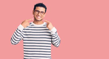 Gündelik kıyafetler ve gözlük takan, neşeli gülümseyen, parmaklarıyla ve ağzıyla işaret eden İspanyol asıllı yakışıklı genç adam. diş sağlığı kavramı. 