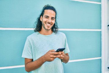 Duvara dayanmış akıllı telefon kullanan, mutlu gülümseyen genç İspanyol adam.