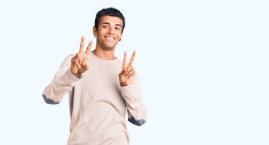 Genç Afrikalı Amerikalı sıradan kıyafetler giymiş gülümseyen ve parmaklarını zafer işareti yaparken gösteren bir adam. İki numara.. 