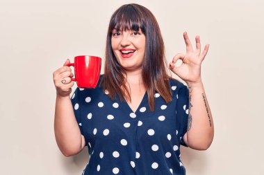 Genç ve cüsseli bir kadın elinde kahve tutuyor. Parmaklarıyla işaret veriyor. Gülümsüyor. 