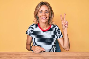 Gündelik giysiler giymiş güzel beyaz kadın masaya oturmuş gülümsüyor ve kameraya göz kırpıyor zafer işareti yapıyor. İki numara.. 