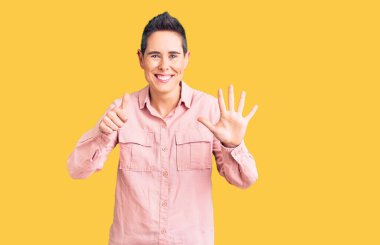 Kısa saçlı, günlük kıyafetler giyen, altı numaralı parmaklarıyla işaret eden, kendinden emin ve mutlu gülümseyen genç bir kadın.. 