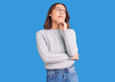 Elleri çenesinde gündelik kıyafetler giyen genç, güzel Çinli kız soru sormayı düşünüyor, düşünceli bir ifade. Gülümseyen ve düşünceli bir yüz. şüphe kavramı. 