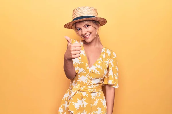 夏の帽子をかぶって休暇中の美しいブロンドの女性と手で幸せな親指アップジェスチャーを行う黄色の背景にドレスアップ 成功を示すカメラを見て表現を承認する — ストック写真