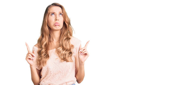 Молодая красивая белая женщина с светлыми волосами в повседневной одежде, выглядящая грустной и расстроенной, указывающая направление пальцами, несчастной и подавленной. 