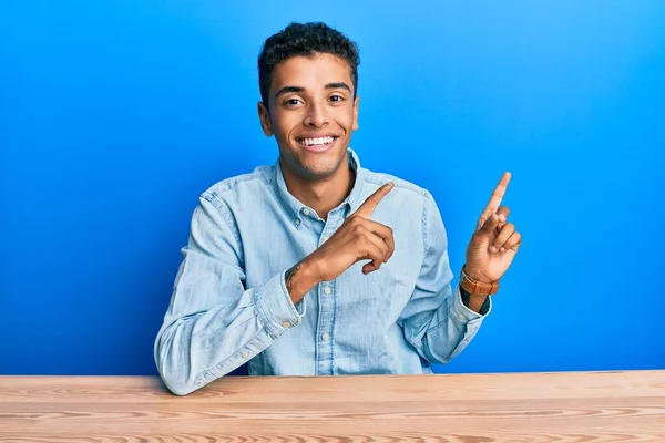 若いですハンサムなアフリカ系アメリカ人男性を着てカジュアルな服をテーブルの上に座っ笑顔とカメラを見て2本の手と指で側に指して — ストック写真