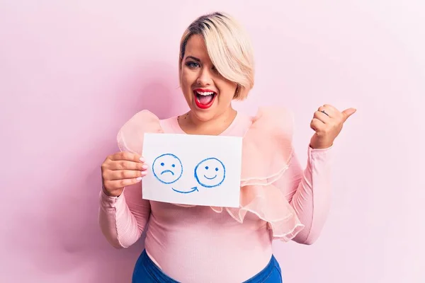 Young blonde plus size woman asking for positive change holding paper with emotion message pointing thumb up to the side smiling happy with open mouth