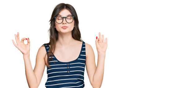 Молодая красивая кавказская девушка в повседневной одежде и очках расслабляется и улыбается с закрытыми глазами, делая медитационный жест пальцами. Концепция йоги. 