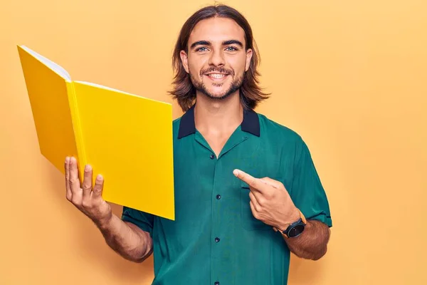 Mladý Pohledný Muž Drží Knihu Úsměvem Šťastný Ukazující Rukou Prstem — Stock fotografie