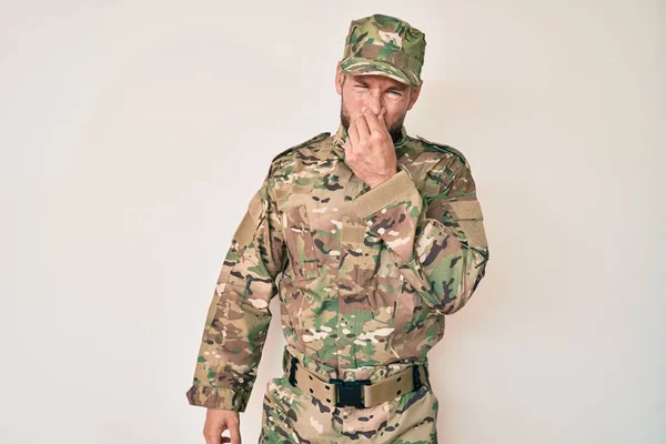 Young caucasian man wearing camouflage army uniform smelling something stinky and disgusting, intolerable smell, holding breath with fingers on nose. bad smell