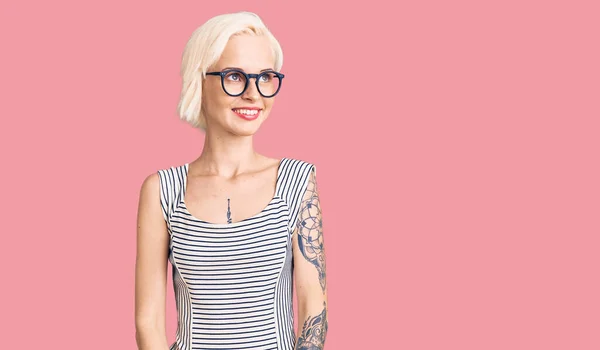Young blonde woman with tattoo wearing casual clothes and glasses looking away to side with smile on face, natural expression. laughing confident.