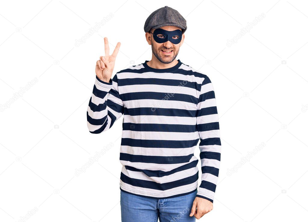 Young handsome man wearing burglar mask smiling looking to the camera showing fingers doing victory sign. number two. 