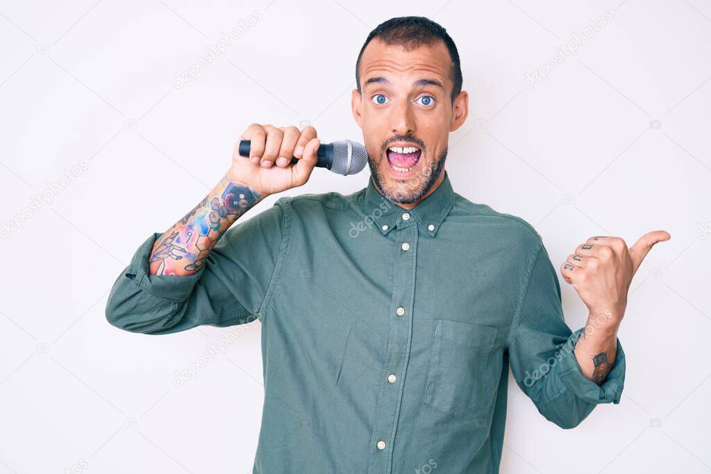 Young handsome man with tattoo singing song using microphone pointing thumb up to the side smiling happy with open mouth 