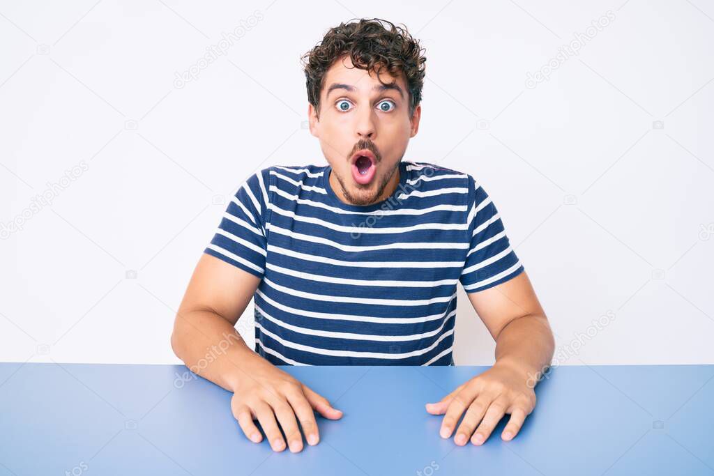 Young caucasian man with curly hair wearing casual clothes sitting on the table scared and amazed with open mouth for surprise, disbelief face 