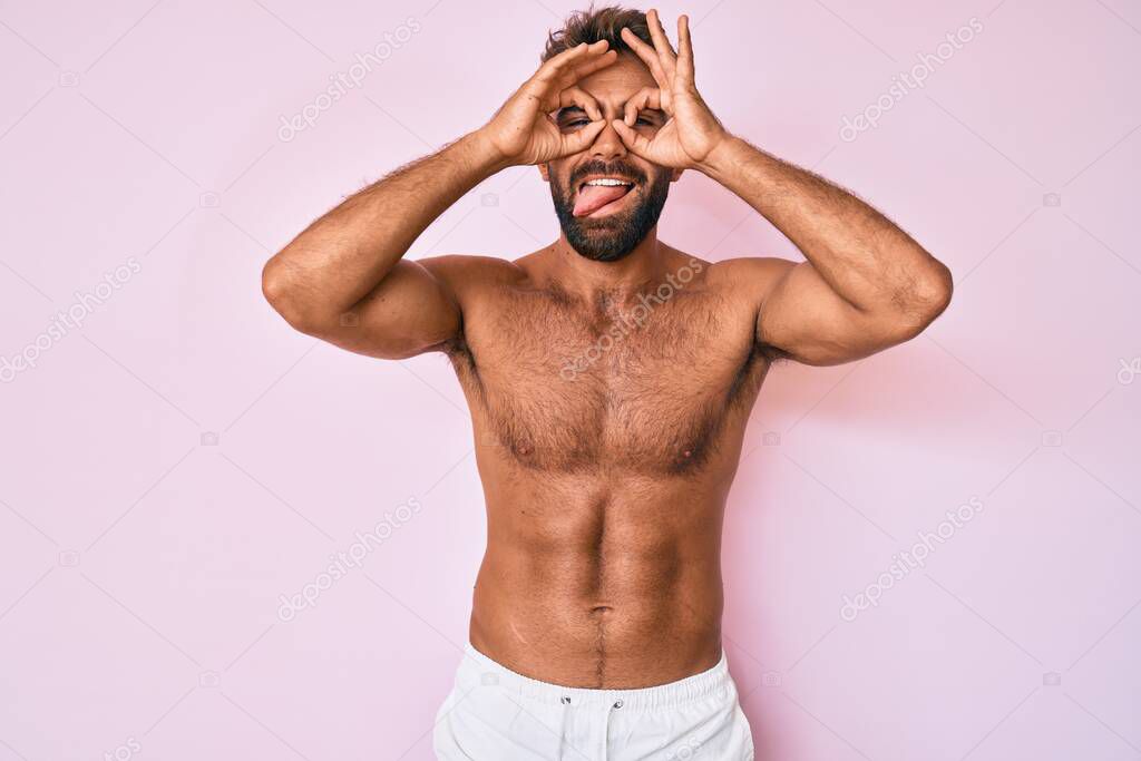 Young hispanic man standing shirtless doing ok gesture like binoculars sticking tongue out, eyes looking through fingers. crazy expression. 