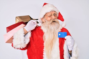 Noel Baba kostümü giymiş yaşlı bir adam elinde alışveriş çantaları ve kredi kartı ile kameraya seksi bir ifadeyle bakıyor, neşeli ve mutlu bir yüzle.. 