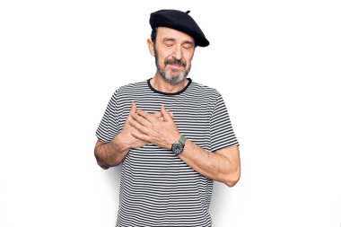 Orta yaşlı yakışıklı bir adam çizgili tişört giyiyor ve beyaz arka planda Fransız beresi takıyor elleri göğsünde gülümsüyor, yüzünde minnet dolu bir jest ile gözleri kapalı. Sağlık kavramı.