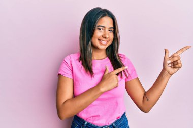 Gündelik kıyafetler giyen, gülümseyen ve kameraya bakan genç Latin kız iki elini ve parmaklarını kenarı işaret ediyor.. 