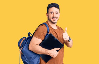 Sırt çantalı genç İspanyol adam elinde bir klasör, gülümsüyor mutlu ve pozitif, başparmağıyla mükemmel ve onay işareti yapıyor. 