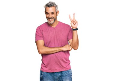 Orta yaşlı, gri saçlı, sıradan kıyafetler giyen, gülümseyen, kameraya göz kırpan, parmaklarıyla zafer işareti yapan bir adam. İki numara.. 