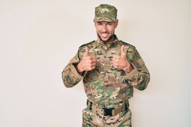 Young caucasian man wearing camouflage army uniform success sign doing positive gesture with hand, thumbs up smiling and happy. cheerful expression and winner gesture.  clipart