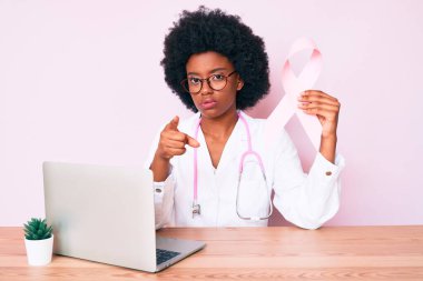 Genç Afro-Amerikan kadın doktor üniforması giyiyor elinde pembe kanser kurdelesi var kamerayı işaret ediyor ve sen kendine güvenen ciddi görünüyorsun. 