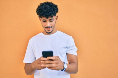 Genç Arap adam mutlu bir şekilde gülümsüyor. Duvara yaslanmış akıllı telefon kullanıyor..