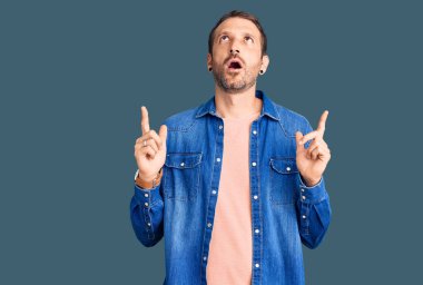 Gündelik giysiler giyen genç yakışıklı adam şaşırmış ve şaşırmış bir şekilde yukarı bakıyor ve parmaklarıyla işaret ediyor ve kollarını kaldırıyor.. 